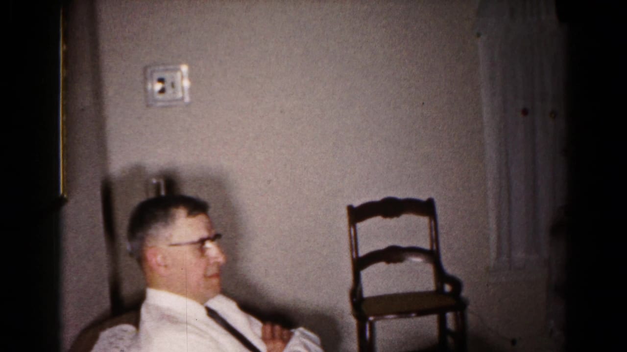 Man in glasses and white shirt seated beside a wall, with wooden chair and window nearby.