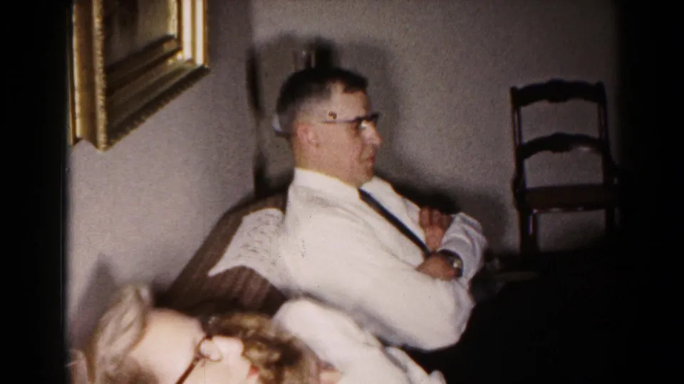 1959 Hagerstown home-movie frame showing a seated man in profile and a woman in glasses leaning through the foreground of a private interior.