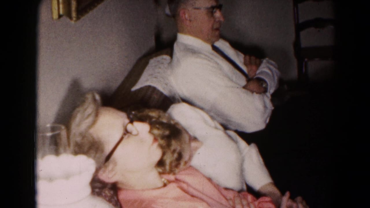 1959 Hagerstown interior frame with a woman in a pink dress in the foreground and a man seated behind her near a wooden chair.