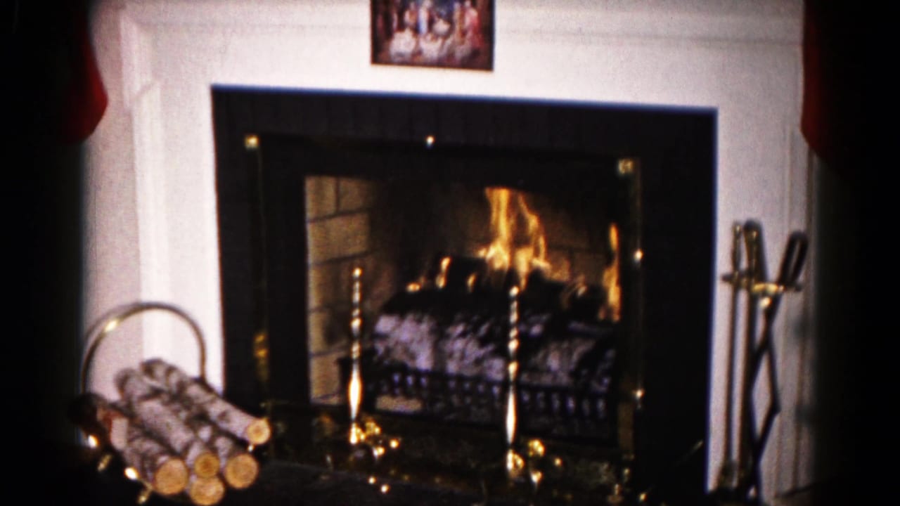 Fireplace with flames, log basket, brass tools, framed picture, and mantel decorations.