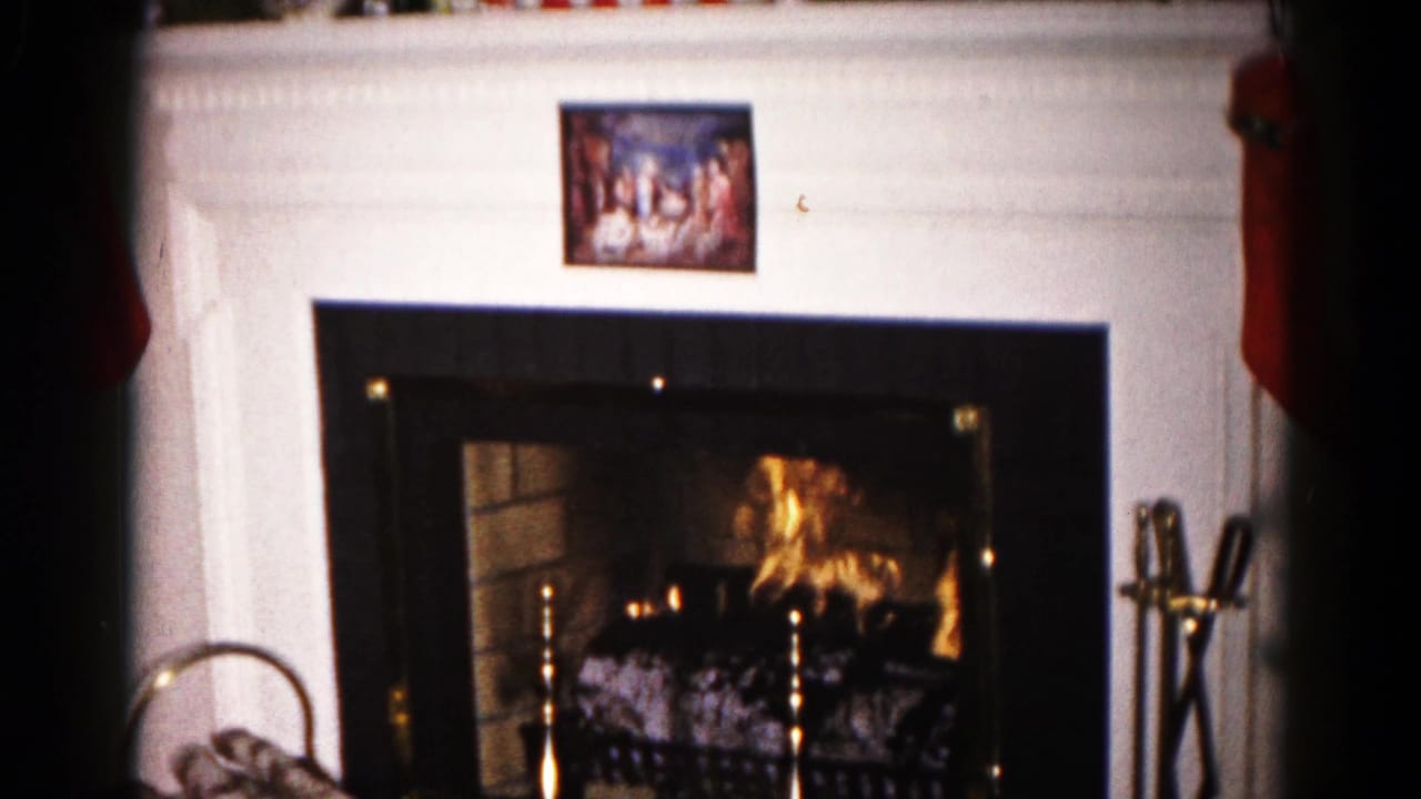 1959 Hagerstown Christmas home-movie frame focused on a lit fireplace below a white mantel and small religious seasonal picture.