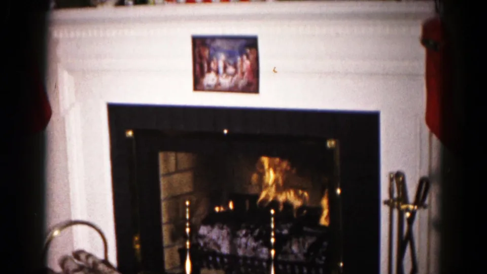 1959 Hagerstown Christmas home-movie frame focused on a lit fireplace below a white mantel and small religious seasonal picture.