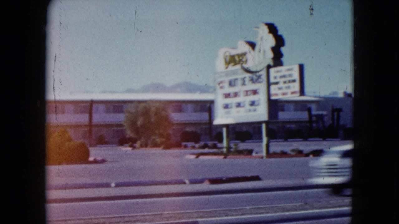 1959 Nevada roadside motel seen from a moving vehicle, with a large sign, parked cars, traffic lanes, and distant mountains.