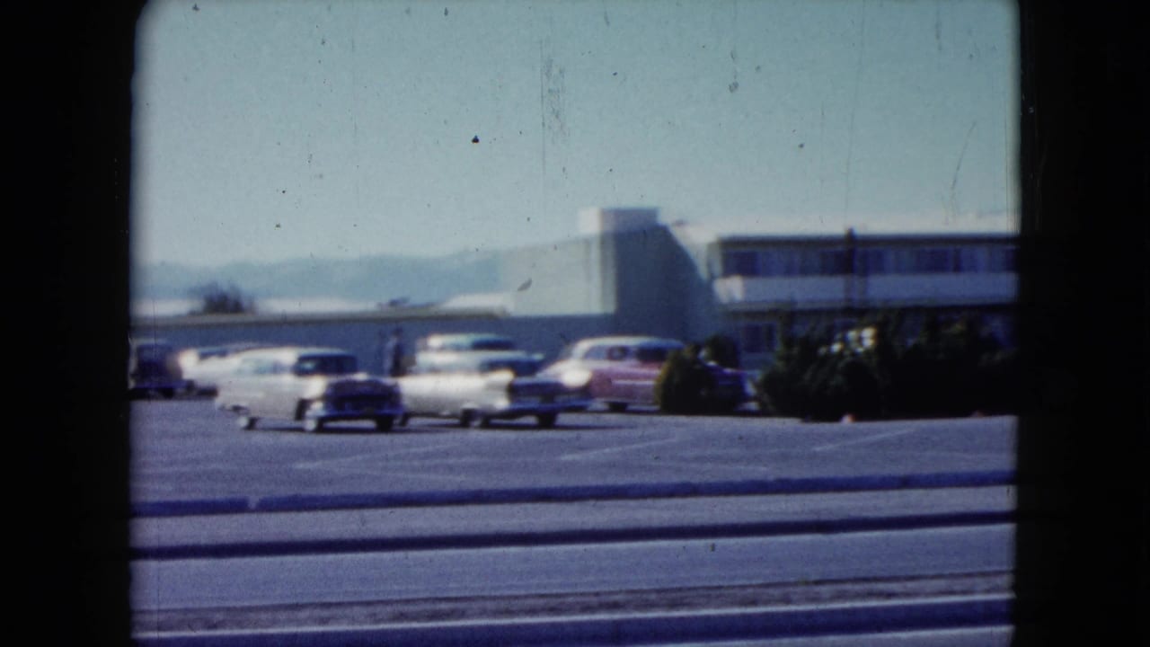 A 1959 Nevada roadside motel lot with several classic cars, low landscaping, curb lines, and the motel building behind them.