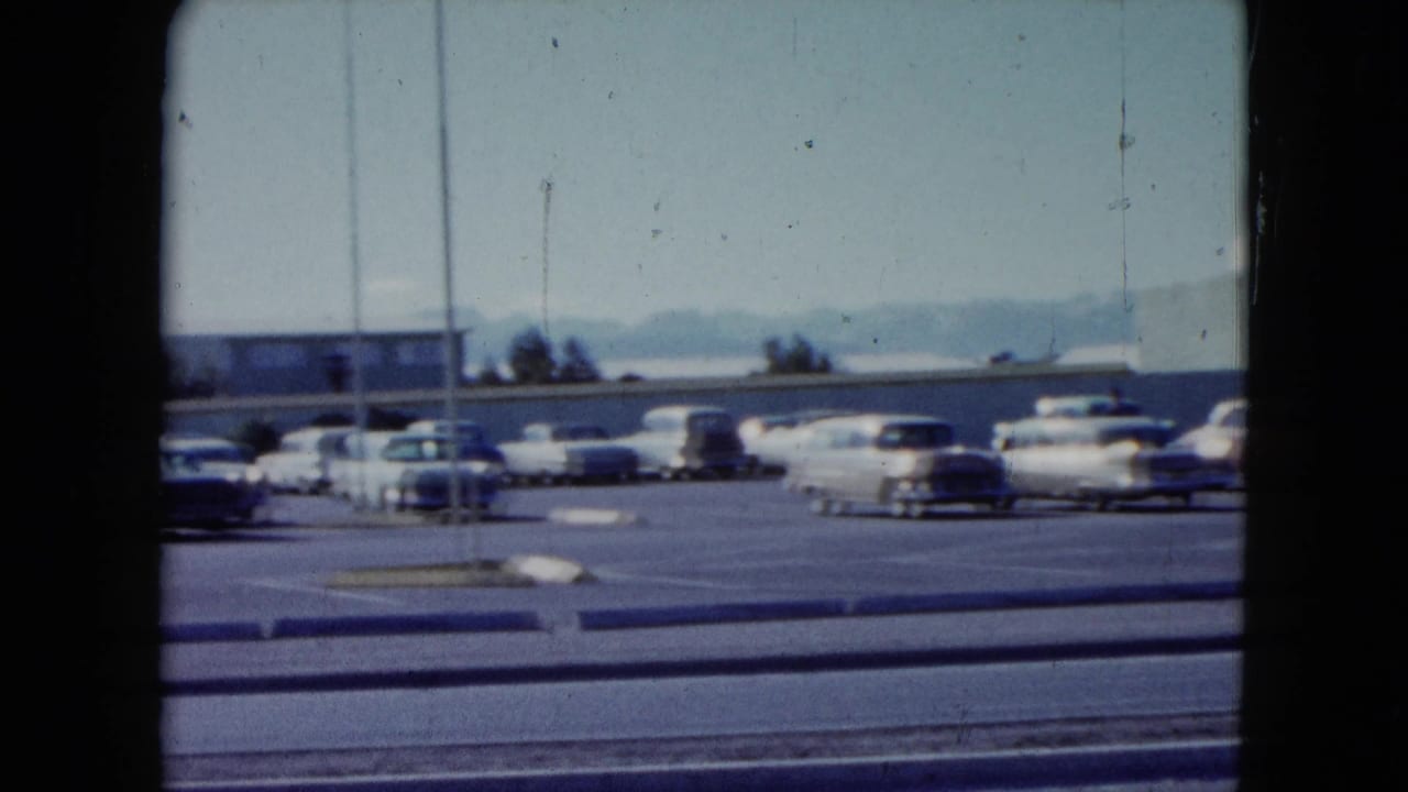 1959 Nevada motel-area parking lot with rows of classic cars, roadside lanes, a light pole, distant mountains, and a clear sky.