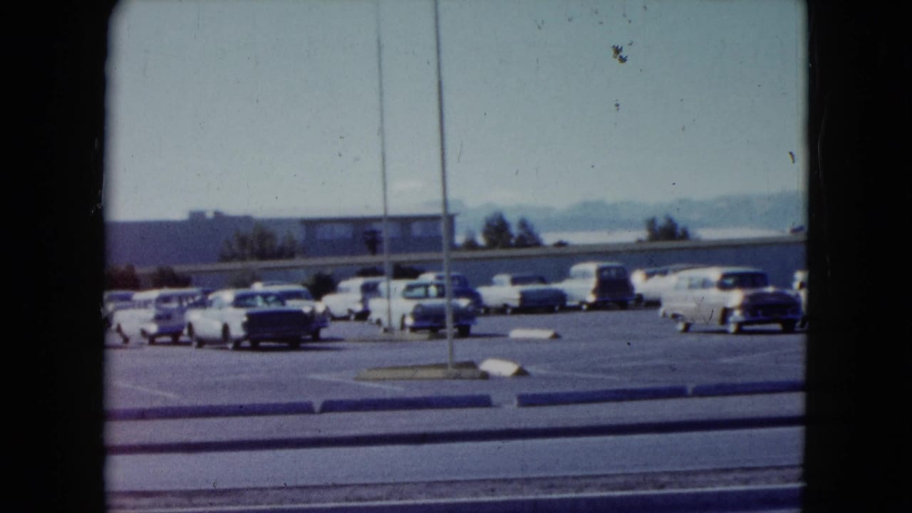 Wide parking lot with late-1950s cars, curb, poles, and low buildings in the distance.