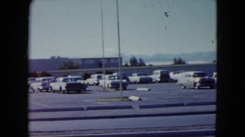 1959 Nevada home-movie frame looking across a sunlit parking lot with period cars, roadside poles, and distant low buildings.