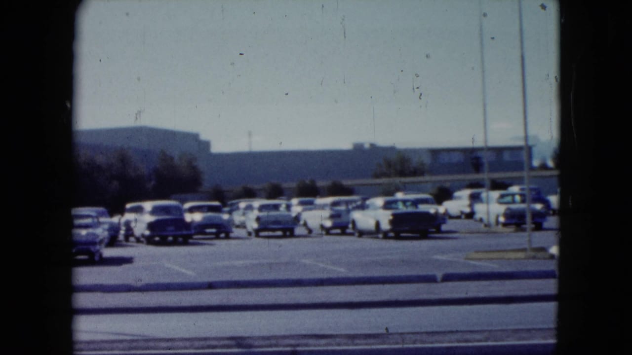 1959 Nevada frame showing a roadside parking lot filled with classic cars, with low buildings and hills beyond.