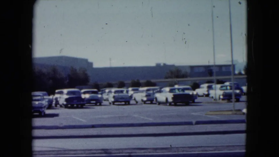 1959 Nevada frame showing a roadside parking lot filled with classic cars, with low buildings and hills beyond.