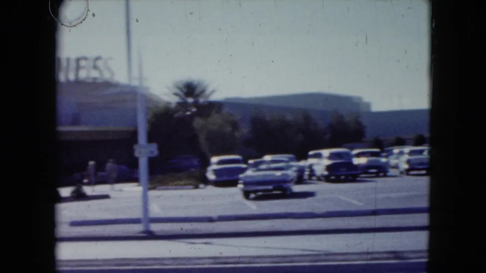 1959 Nevada home-movie frame passing the Dunes frontage, with classic cars, a palm tree, pole, and hills visible.