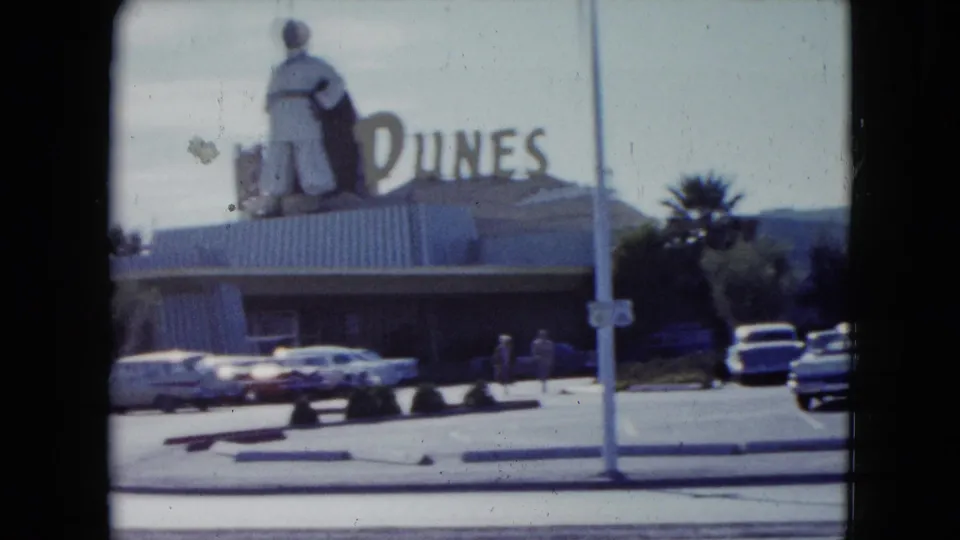 1959 Nevada frame showing the Dunes sign and rooftop figure above a low building, with cars and small pedestrians nearby.