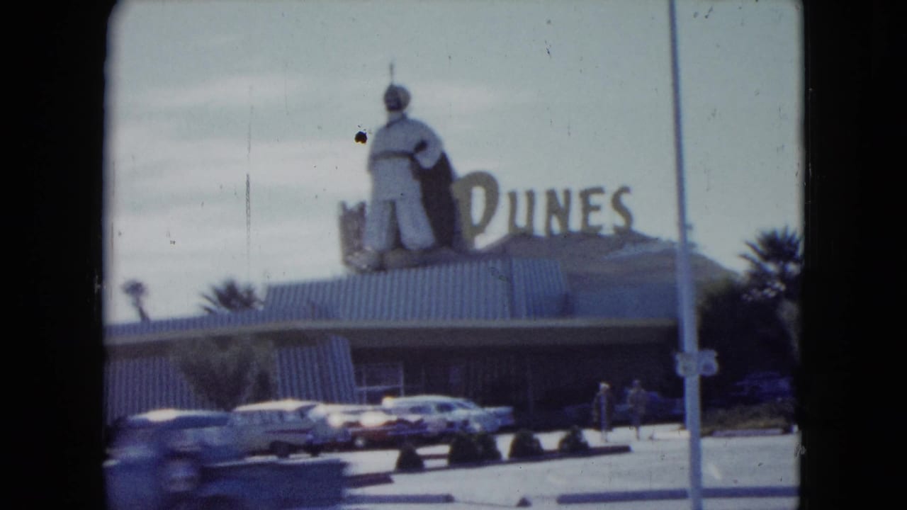 1959 Nevada home-movie frame showing the Dunes hotel facade, rooftop figure, parked classic cars, palm trees, and pedestrians.