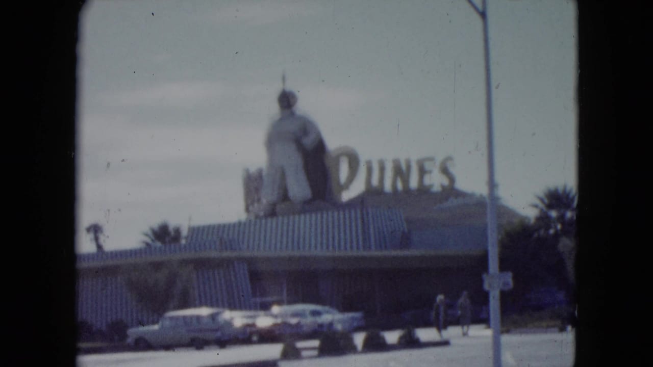 1959 Nevada frame with the Dunes sign, large rooftop figure, parked period cars, street pole, palms, and pedestrians.