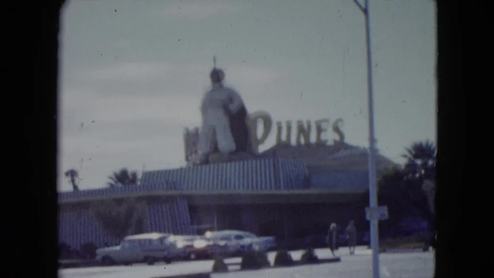 1959 Nevada frame with the Dunes sign, large rooftop figure, parked period cars, street pole, palms, and pedestrians.