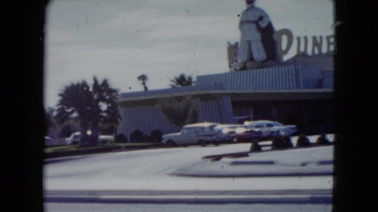 1959 Nevada home-movie view of the Dunes facade, cropped rooftop figure, parked classic cars, palms, curb, and broad street foreground.