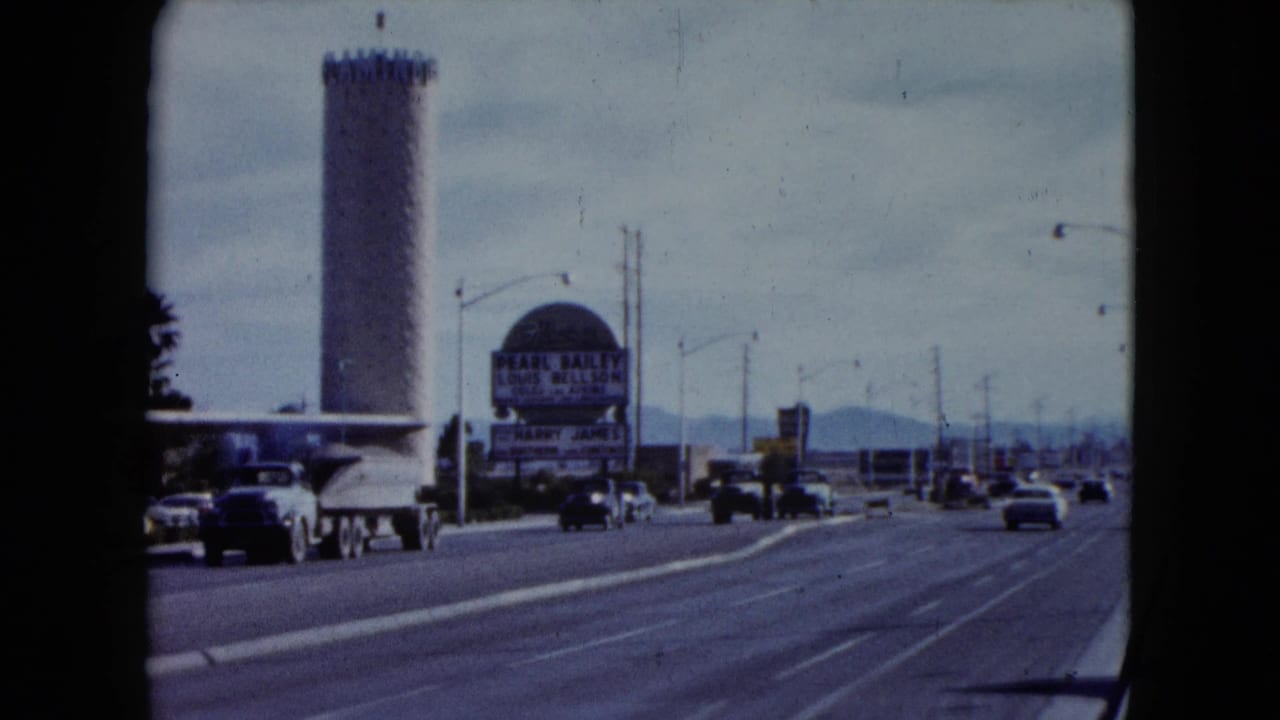 1959 Nevada street view showing a tall resort tower, roadside hotel sign, moving and parked cars, streetlights, and distant mountains.