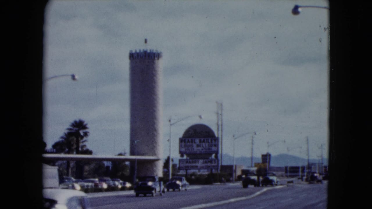 Cars pass a roadside tower and large sign on a sunny multi-lane street.