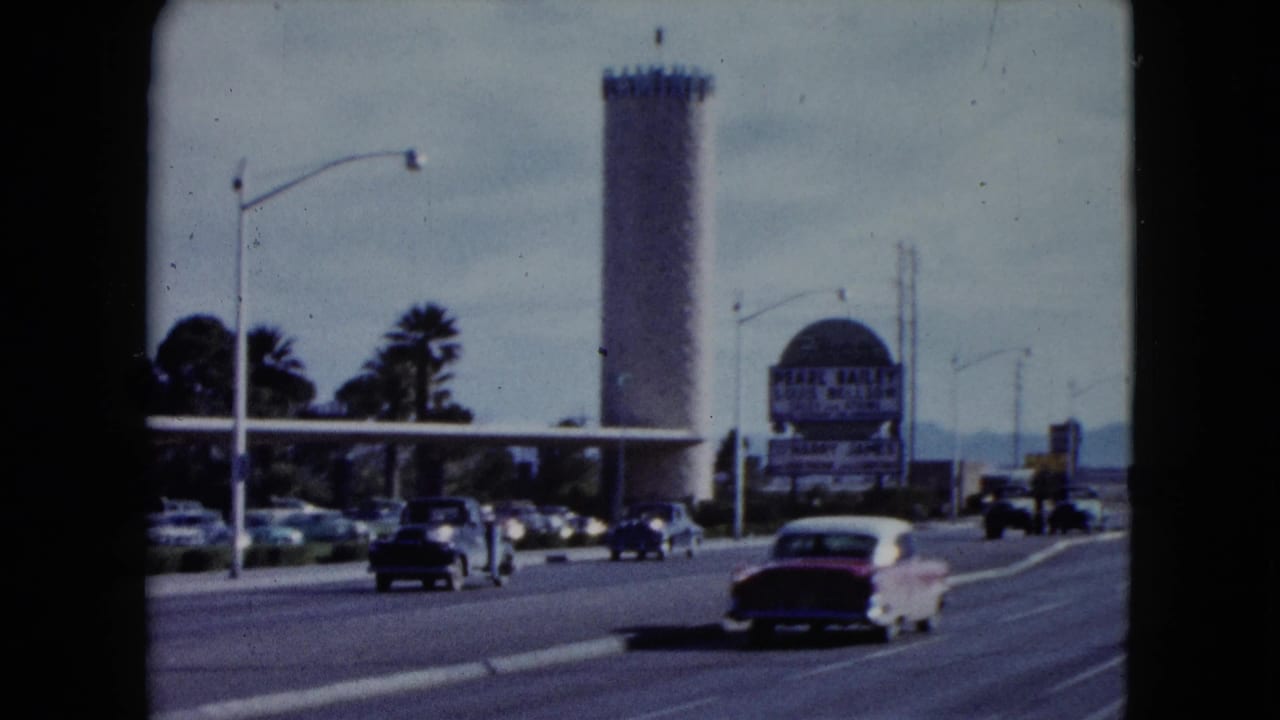 1959 Nevada street view with several classic cars, a tan cylindrical tower, palm trees, streetlights, and entertainment signage beside the road.