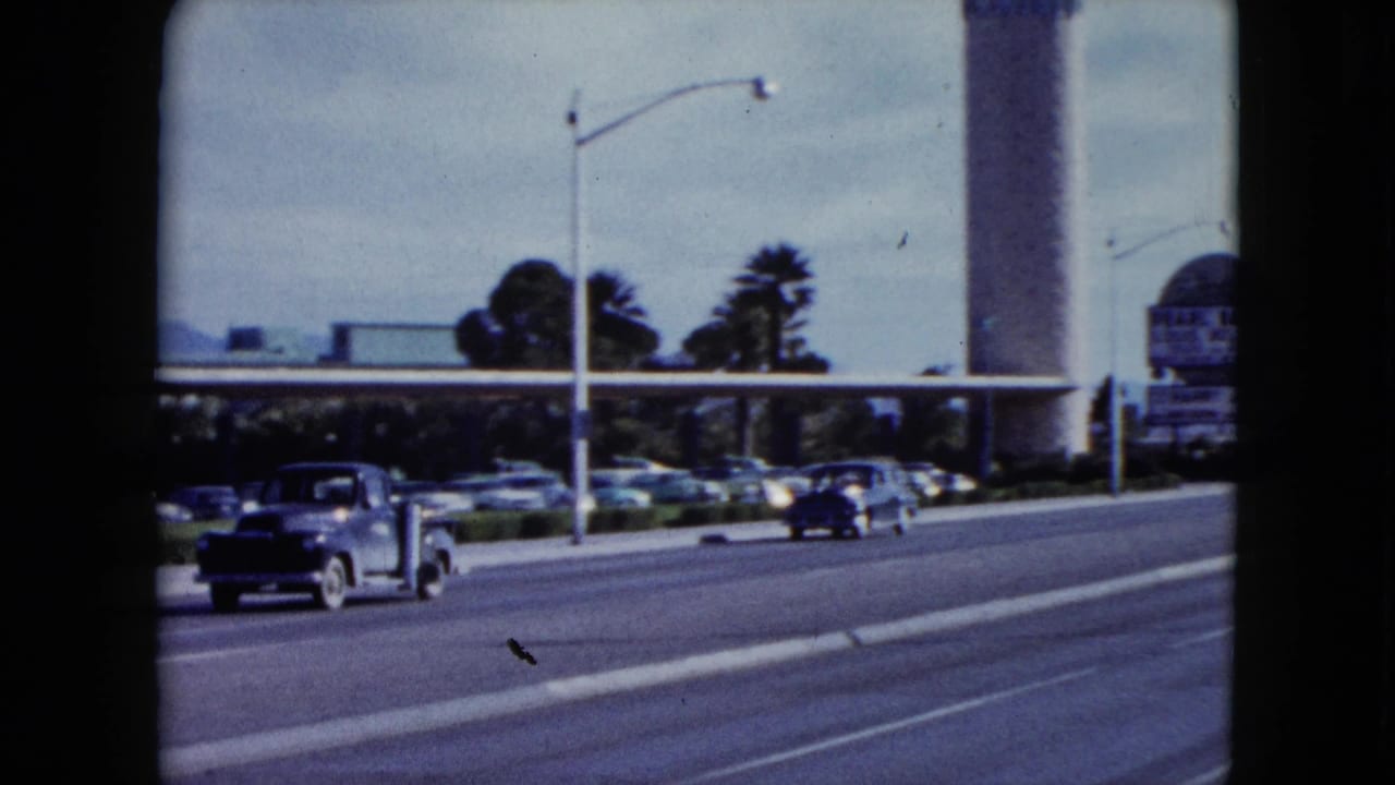 Nevada 8mm frame from 1959 showing cars passing a low canopy and palm trees with the tan tower and partial roadside sign at the edge.