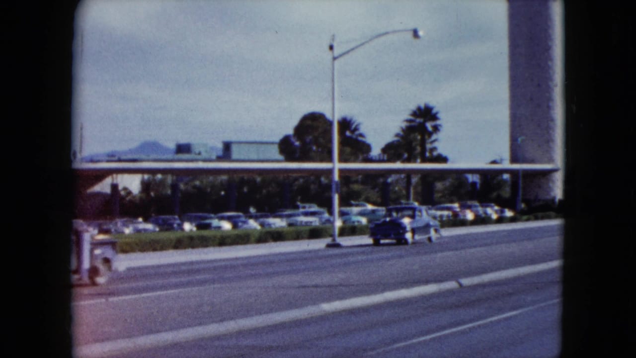 1959 Nevada 8mm frame showing a classic car on a multi-lane road near parked autos, palm trees, and a large roadside building.