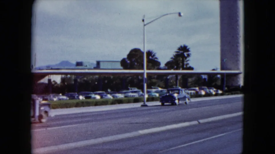1959 Nevada 8mm frame showing a classic car on a multi-lane road near parked autos, palm trees, and a large roadside building.