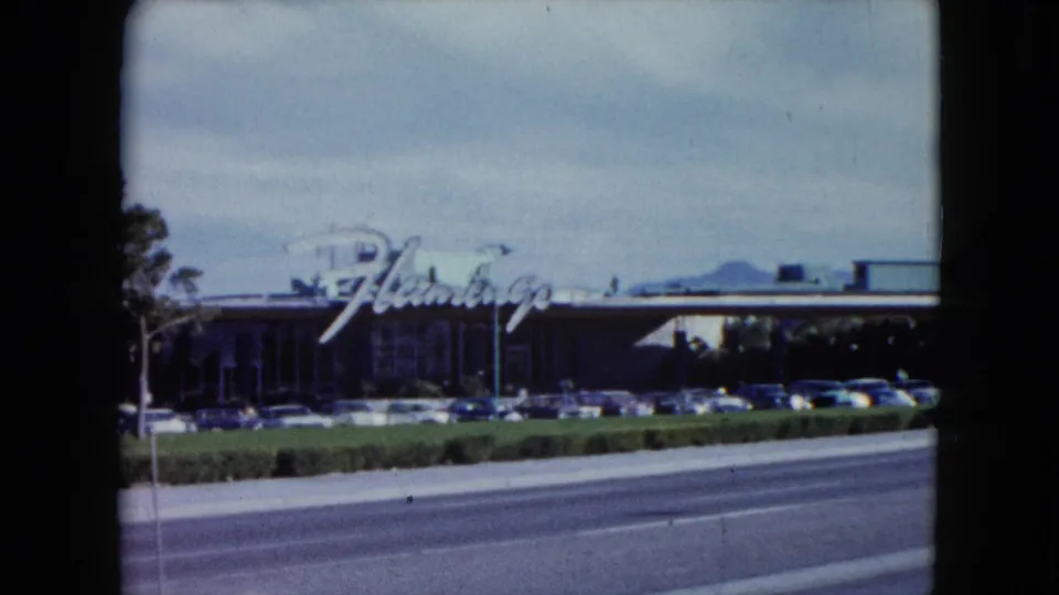 1959 Nevada 8mm frame showing the Flamingo sign, a long roadside building, vintage parked cars, and a broad road in front.