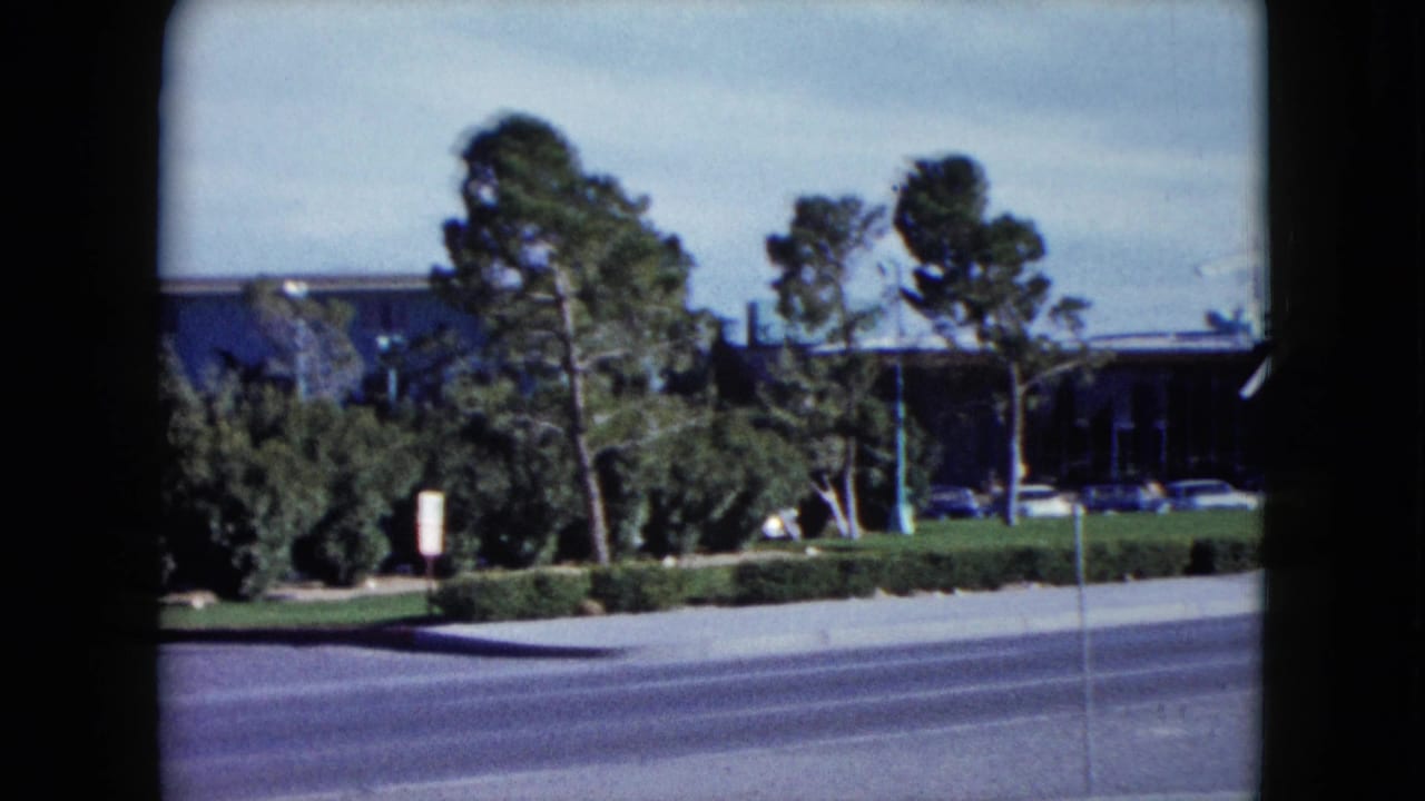 1959 8mm roadside frame showing the Flamingo Hotel area partly obscured by trees and landscaping along a Las Vegas street.