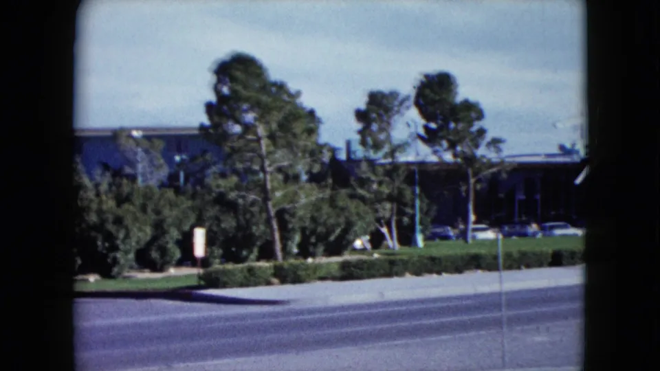 1959 8mm roadside frame showing the Flamingo Hotel area partly obscured by trees and landscaping along a Las Vegas street.