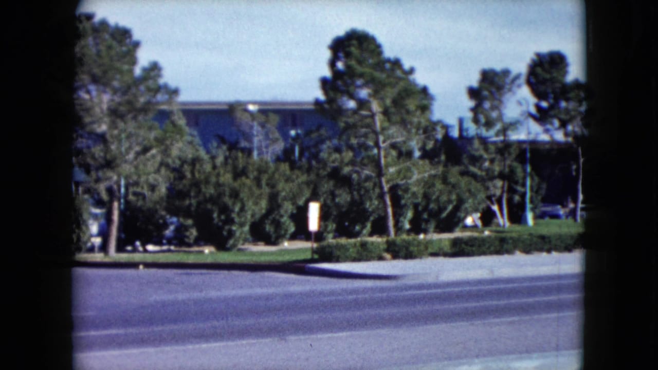 Final 1959 home-movie frame of the Las Vegas hotel exterior, emphasizing roadside landscaping, trees, and parked cars.