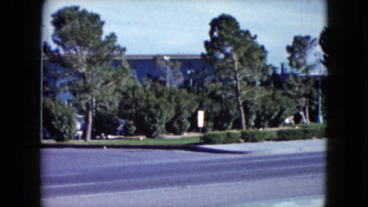 1959 Nevada 8mm frame showing a broad street, hedges, tall trees, and a blue mid-century building in the background.