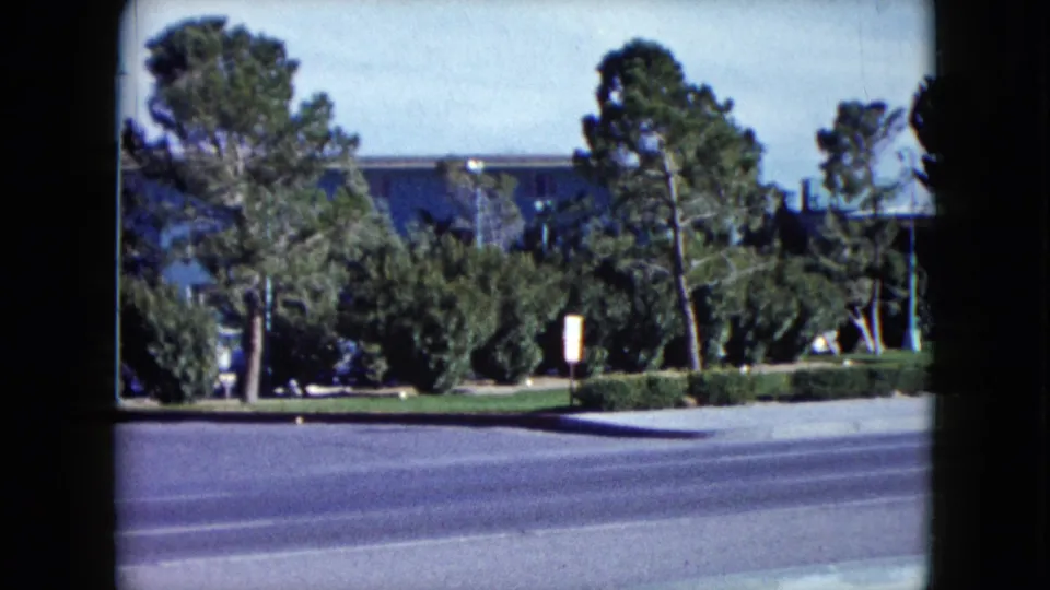 1959 Nevada 8mm frame showing a broad street, hedges, tall trees, and a blue mid-century building in the background.