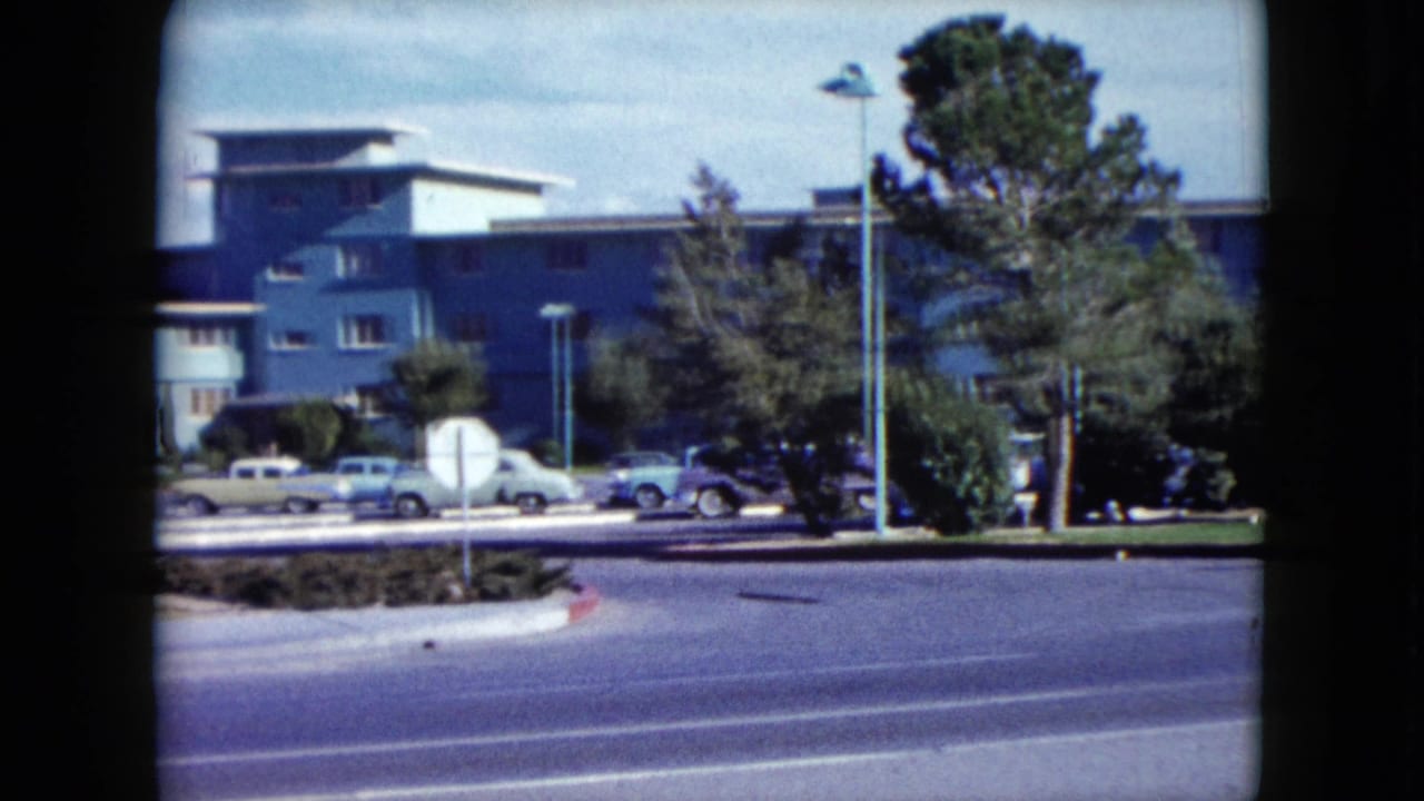 1959 Nevada frame showing a blue mid-century building, classic parked cars, street lamps, trees, and a wide foreground road.