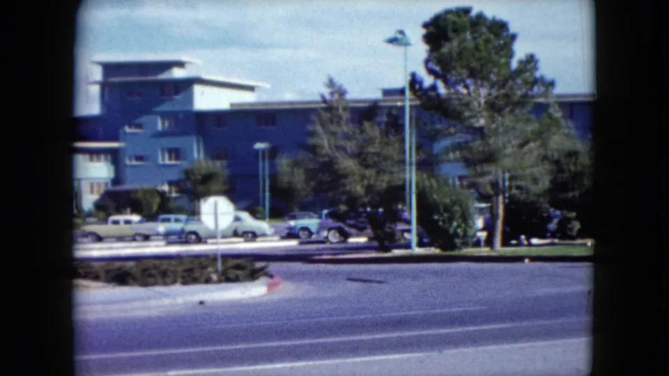 1959 Nevada frame showing a blue mid-century building, classic parked cars, street lamps, trees, and a wide foreground road.