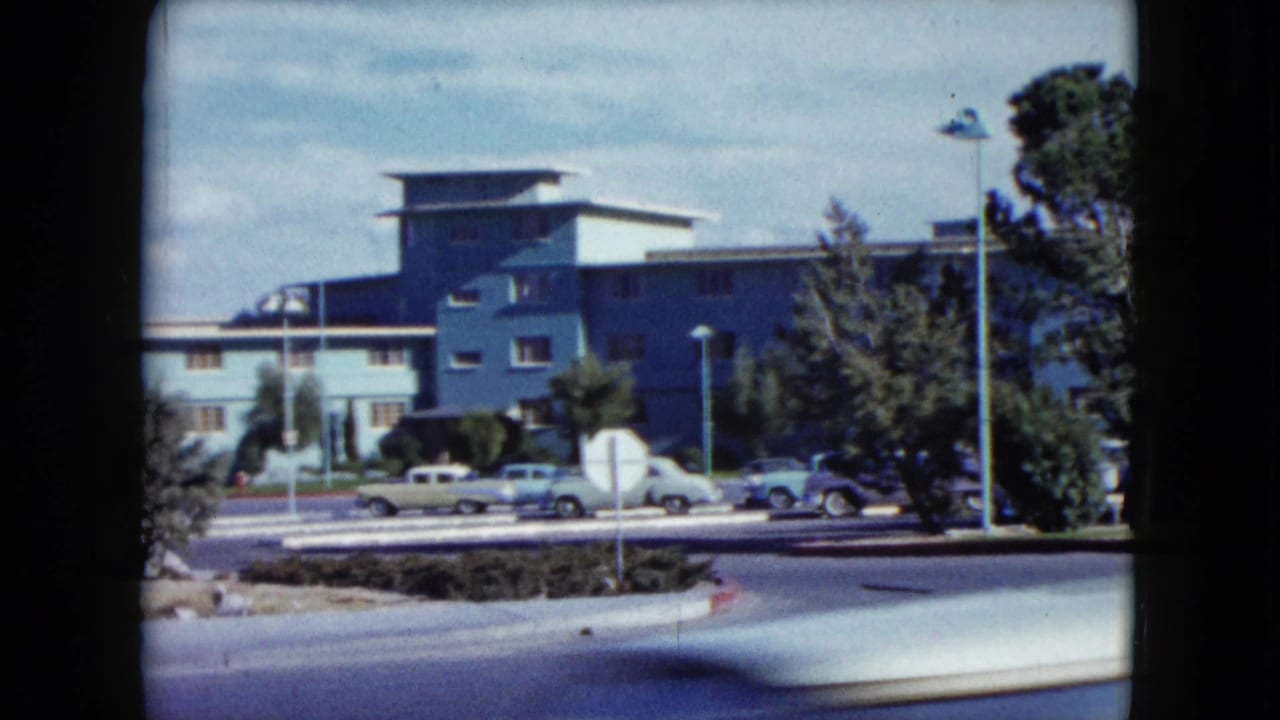 1959 Nevada 8mm frame with a blue building, parking lot, classic cars, trees, and street lamps seen from a moving vehicle.