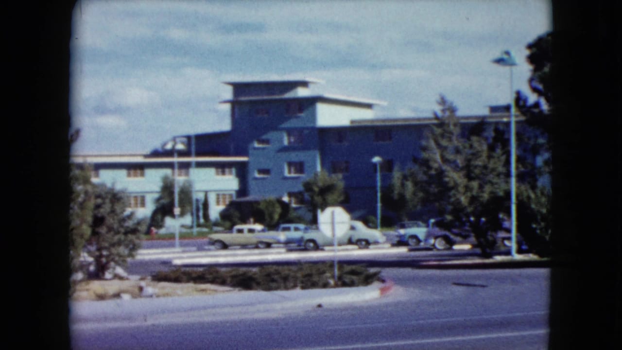 1959 Nevada home-movie frame showing a street, parked cars, trees, and a modern hotel-like building.