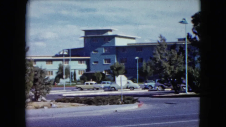 1959 Nevada home-movie frame showing a street, parked cars, trees, and a modern hotel-like building.