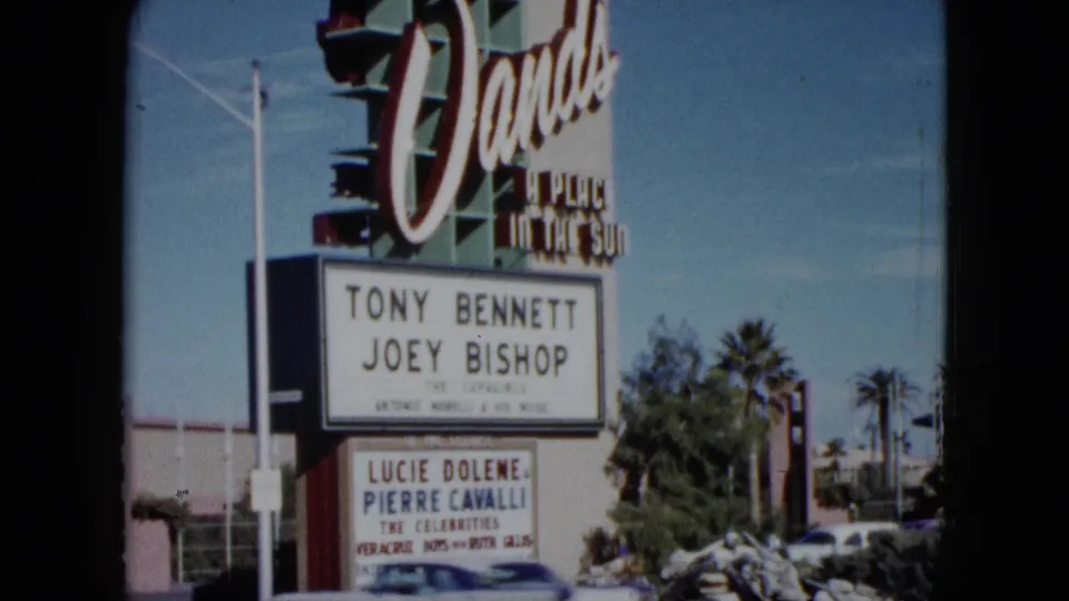 1959 Nevada frame showing the Dunes sign, performer names on the marquee, palm trees, and roadside landscaping.