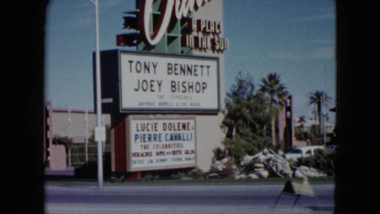 1959 Nevada home-movie frame with the Dunes Hotel marquee, lower performer board, road edge, and desert-resort landscaping.