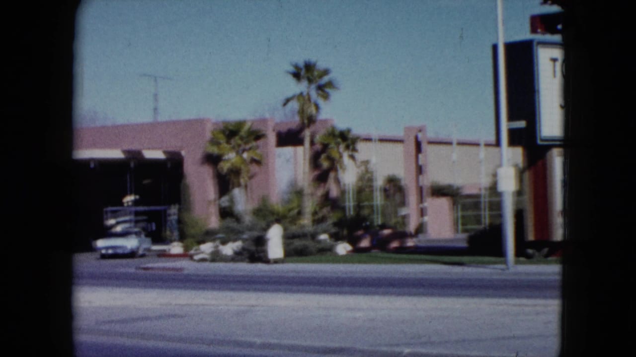 1959 Nevada frame with a venue entrance, palm trees, a parked car, and a robed pedestrian near the sidewalk.