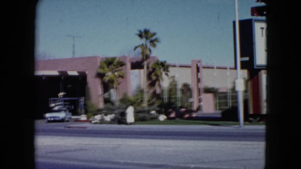 1959 Nevada frame with a venue entrance, palm trees, a parked car, and a robed pedestrian near the sidewalk.