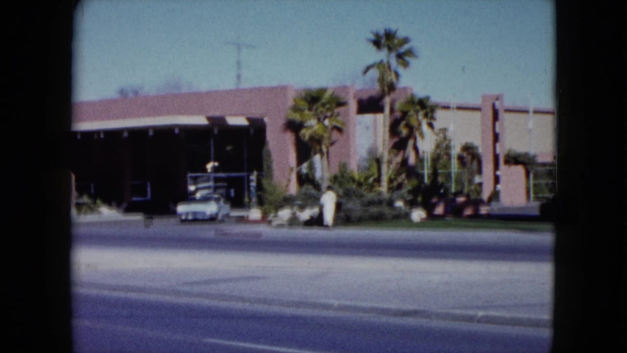 1959 Nevada 8mm frame showing the venue frontage, palm trees, an older car, and a person in a white robe walking nearby.