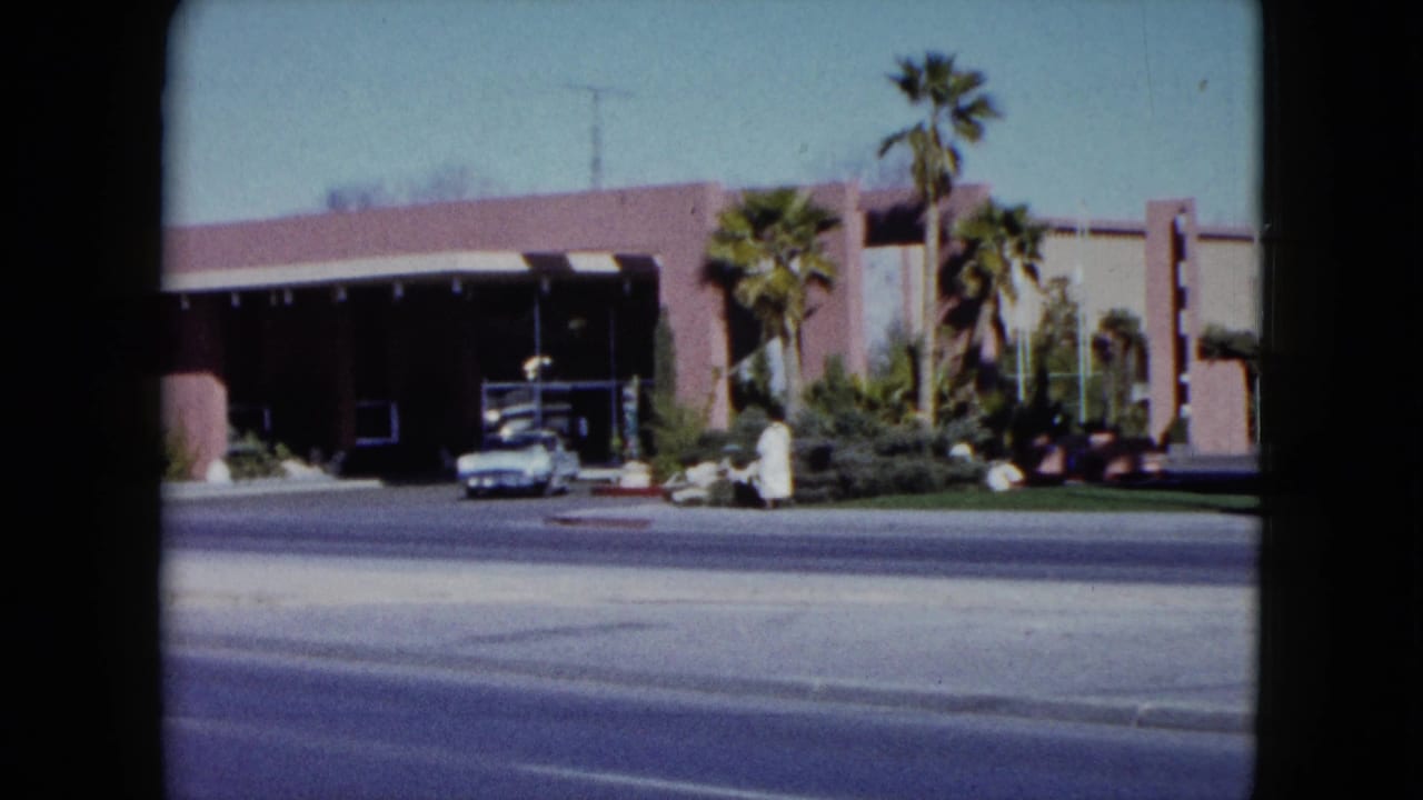 1959 Nevada 8mm frame showing a pink roadside building with palm trees, a covered entrance, a white car, and a person in light clothing.