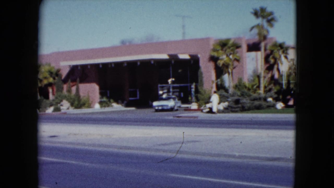 Pink building across a wide street with palm trees, shrubs, a white car, and a person walking nearby.