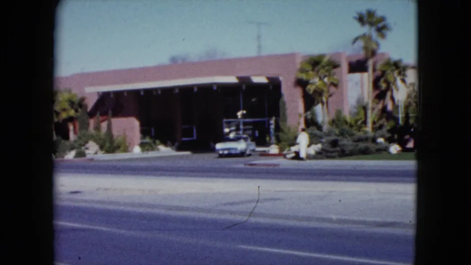 1959 Nevada home-movie frame of a pink building across a sunny street, with palms, landscaping, a white parked car, and a visible pedestrian.