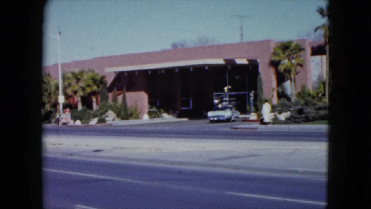 1959 Nevada 8mm frame with a pink commercial-looking building, palms, a shaded entrance, a white car, rocks, shrubs, and road surface.