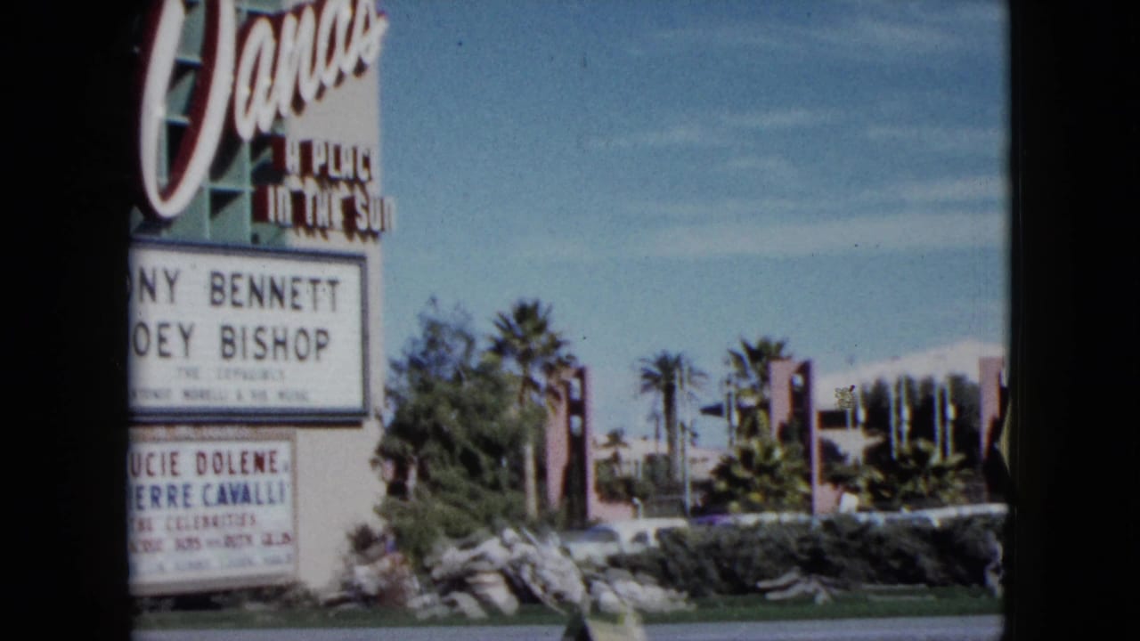 1959 Nevada frame showing a roadside signboard with readable names, palms, landscaped rocks, blue sky, and part of a pink building exterior.
