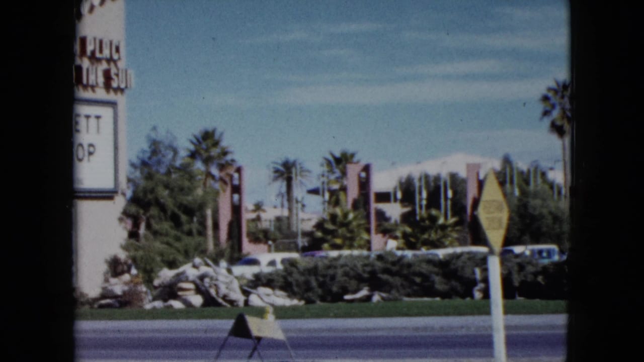 1959 Nevada 8mm frame showing a sunlit roadside with palms, parked cars, landscaping, a yellow road sign, and partial signage at the edge.
