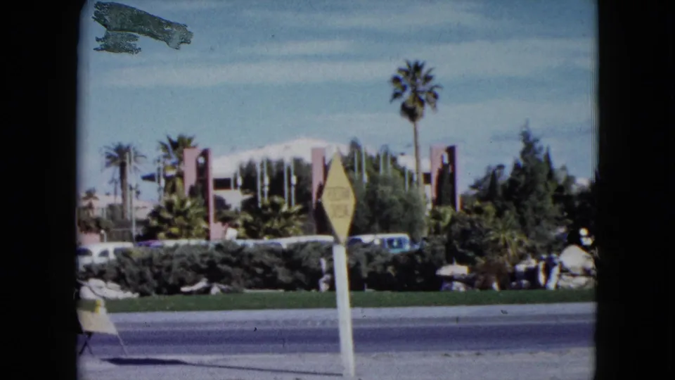 1959 Nevada roadside frame with palm trees, a yellow diamond sign, parked cars, hedges, rock landscaping, and a low modern building.