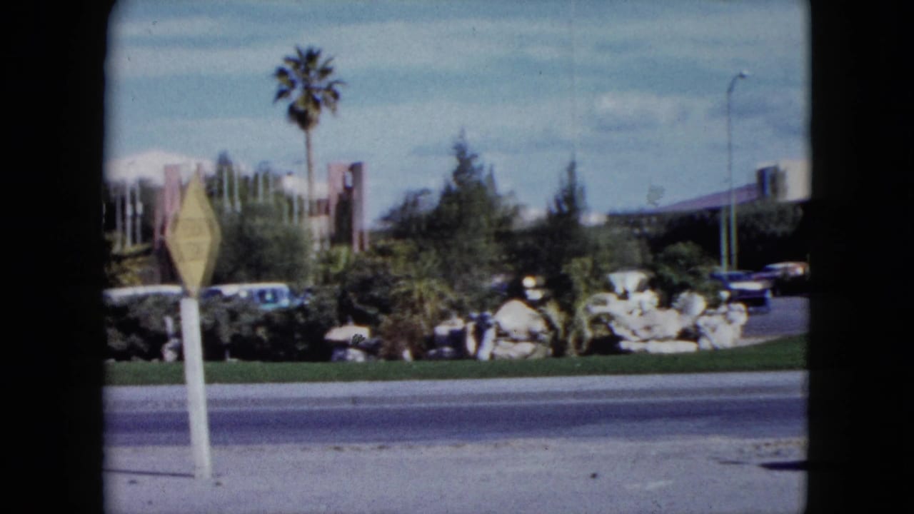 1959 Nevada 8mm view with a yellow road sign in the foreground, palm landscaping, parked cars, rocks, shrubs, and roadside architecture.