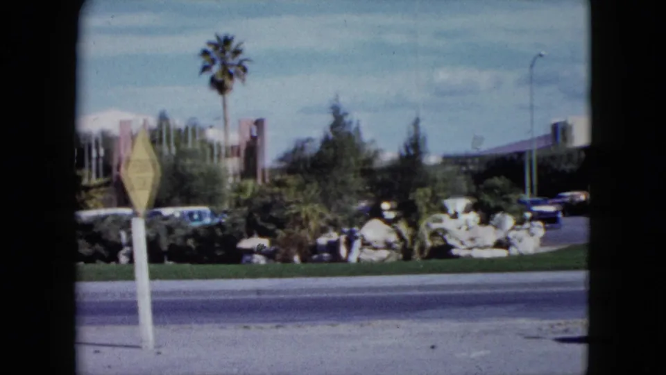 1959 Nevada 8mm view with a yellow road sign in the foreground, palm landscaping, parked cars, rocks, shrubs, and roadside architecture.